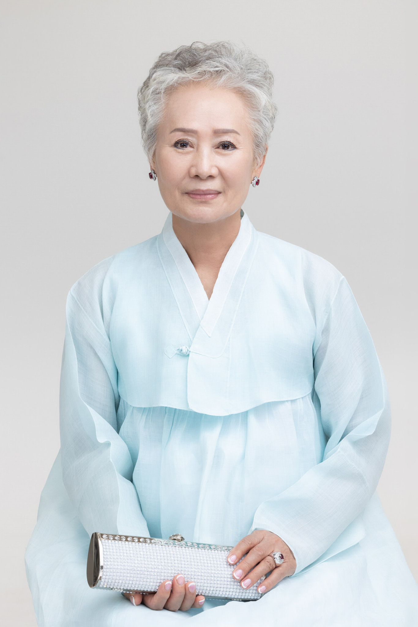 Person with short grey hair wearing a pale blue hanbok and holding a silver clutch against a light grey background.