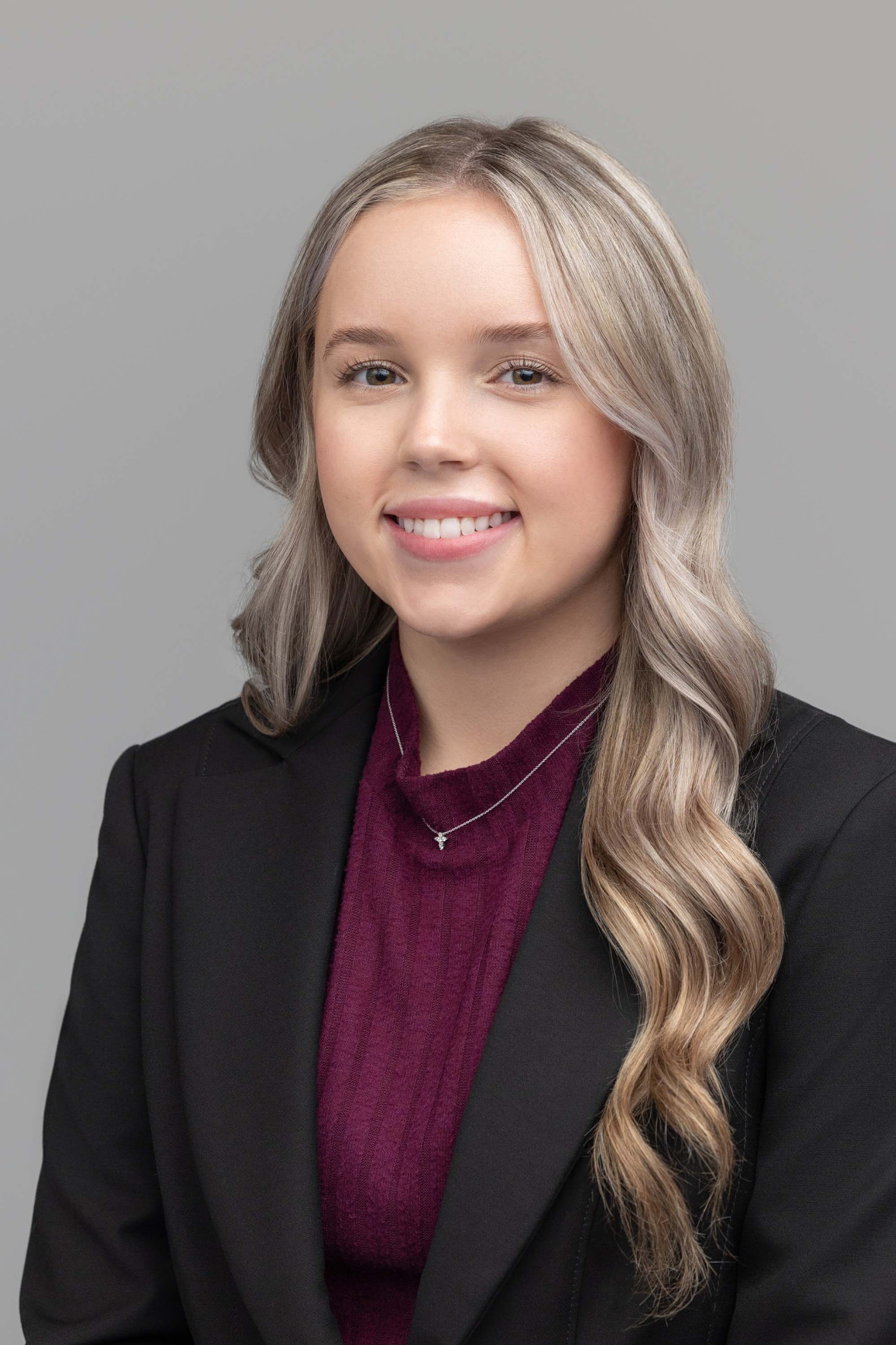 Person with long wavy blonde hair wearing a black blazer, burgundy top and silver necklace against a grey background.