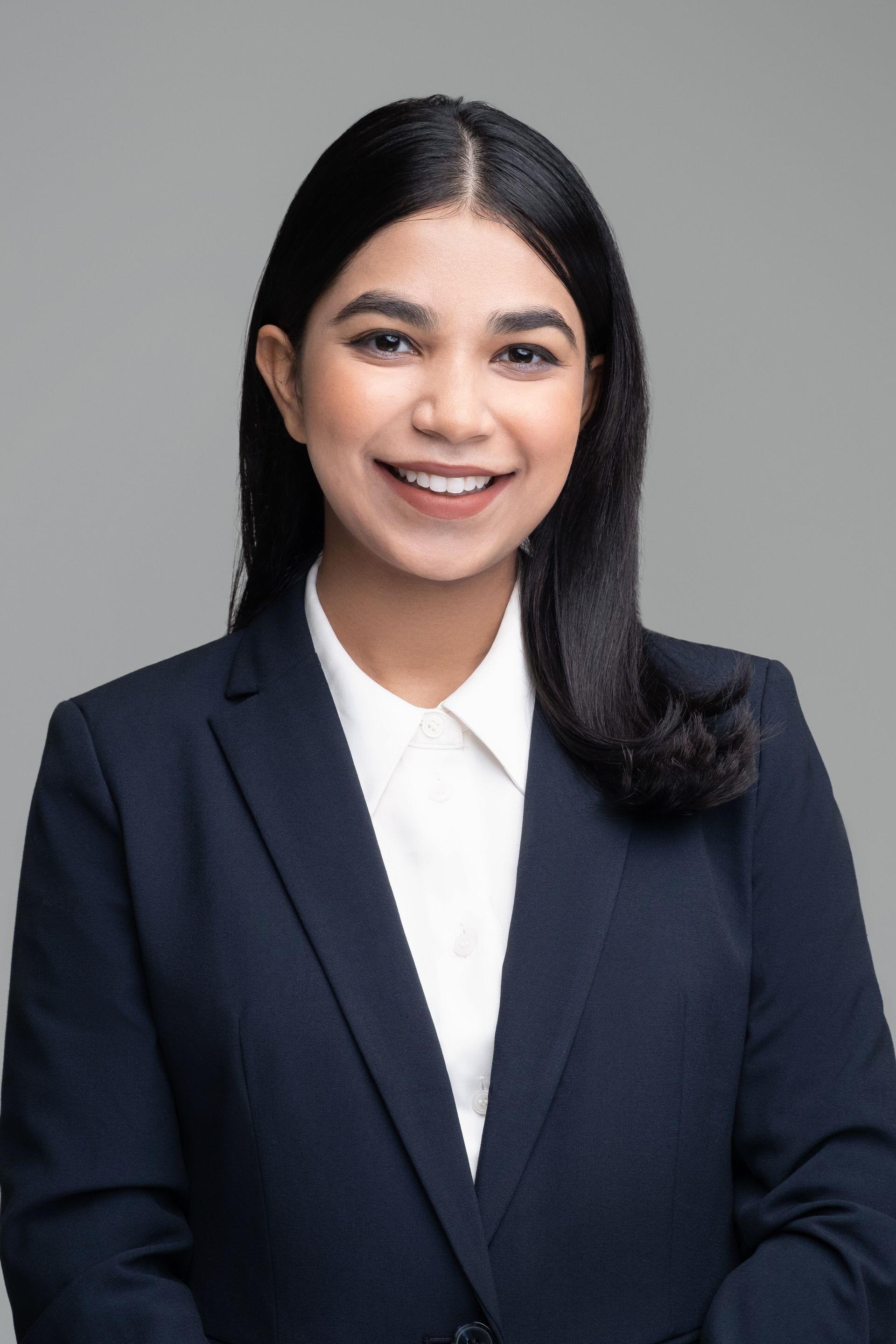 Person with long dark hair wearing a navy jacket and white shirt against a light grey background.