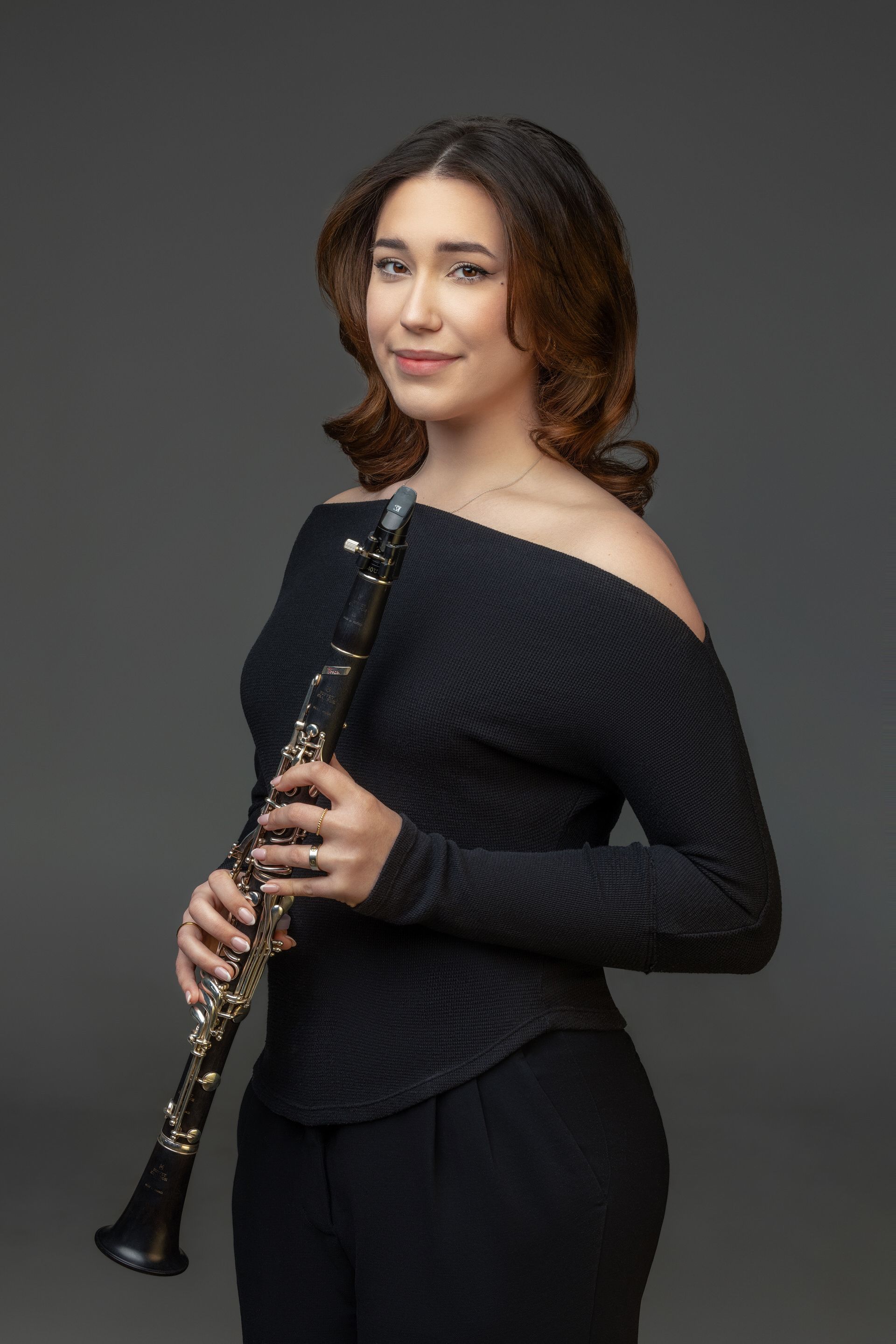 Person wearing a black off-the-shoulder top and holding a clarinet against a grey background.