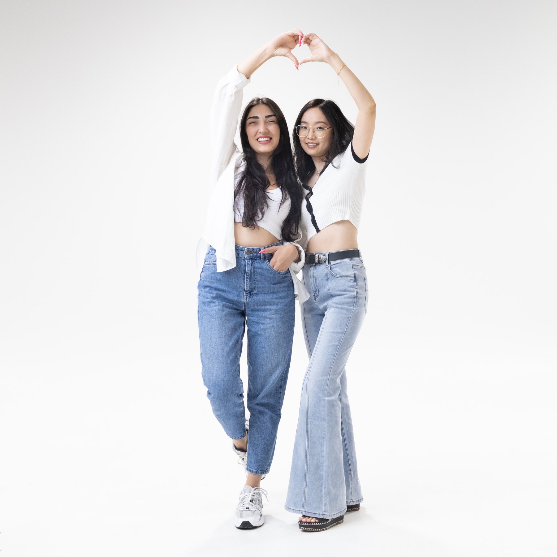 Two smiling people in white tops and jeans forming a heart overhead with their arms.