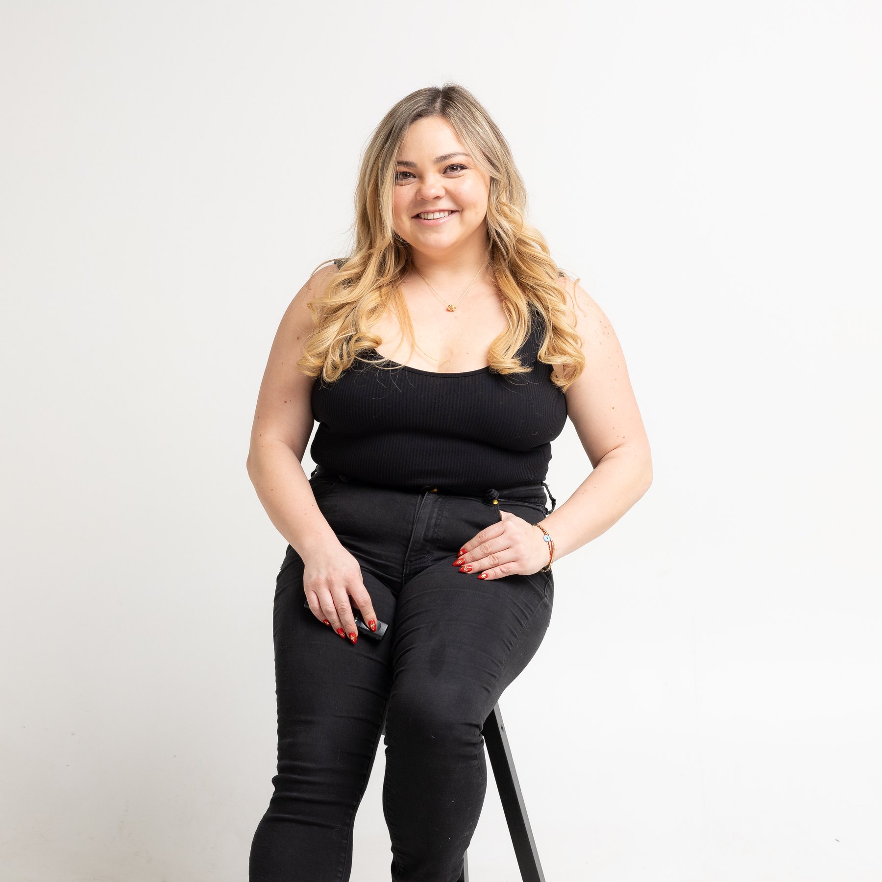Smiling person dressed in black, seated on a stool with a remote in hand.