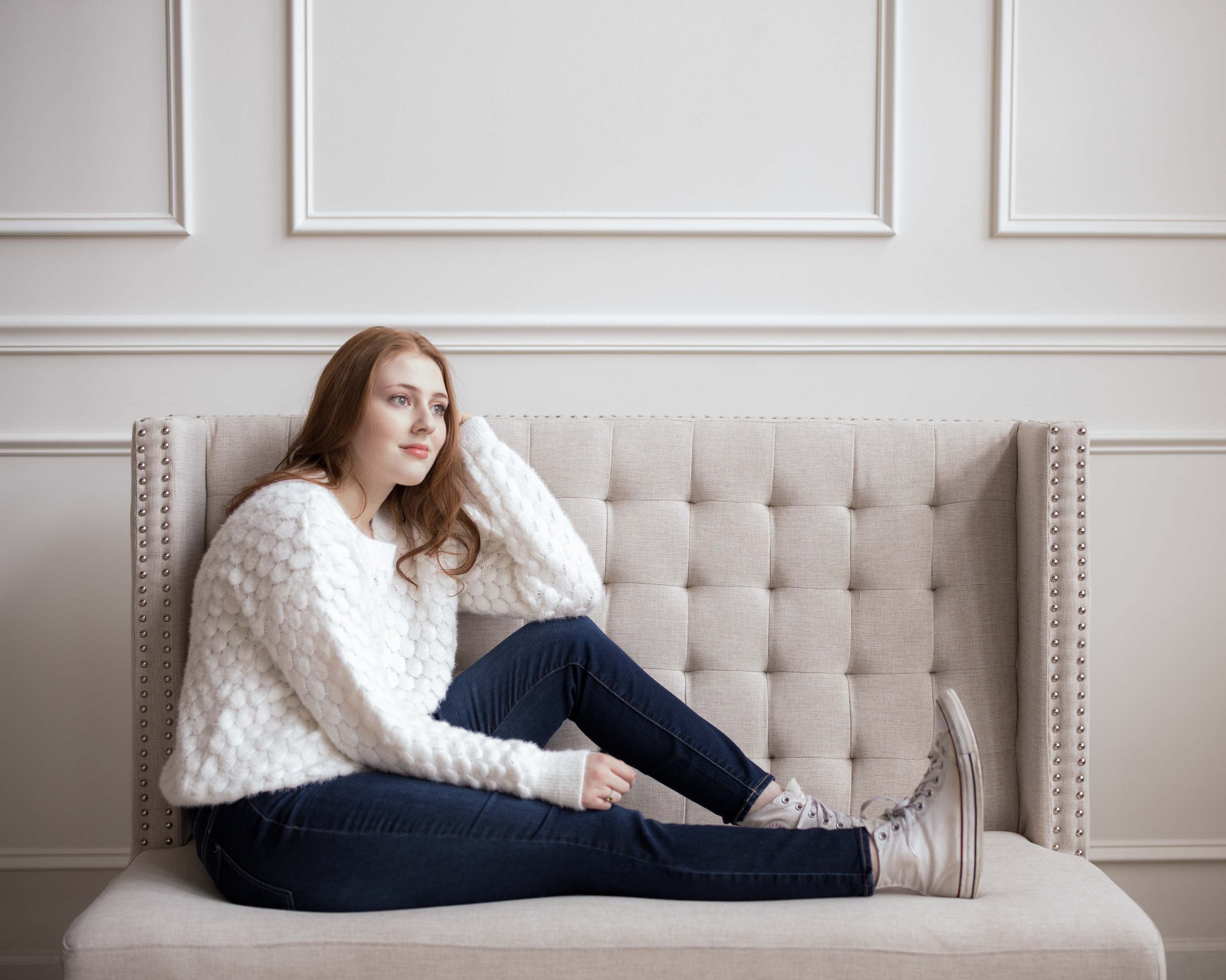 Person in a white sweater sitting sideways on a beige sofa.